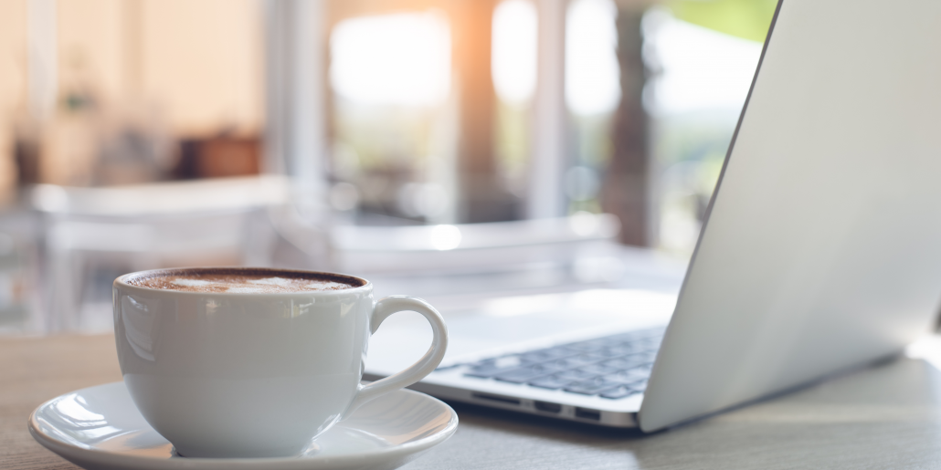 A cup of tea sits in a saucer next to an open lap top. There is a window in the background