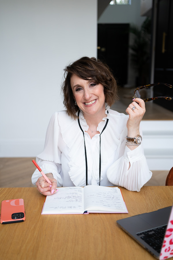 Dani Diosi wearing a white blouse with a black tie holding a pair of glasses and smiling at the camera whilst embarking on a hypnobirthing practitioner training course