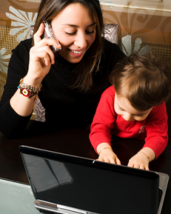 Young mother balancing her baby on her lap who is trying to type on her laptop keyboard. The mother is on the phone