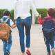 A mother accompanies two children into school. She is wearing a blouse and jeans and they both have backpacks on their backs