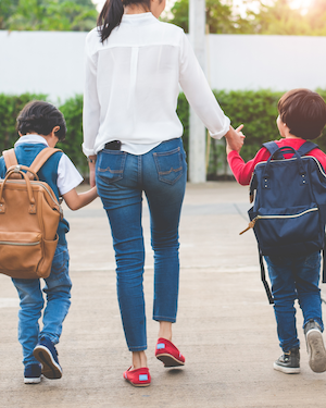 A mother accompanies two children into school. She is wearing a blouse and jeans and they both have backpacks on their backs