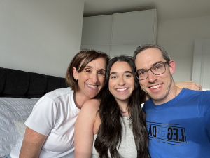 A young girl is surrounded on both sides by her parents. They are all sitting on her bed and they are smiling at the camera