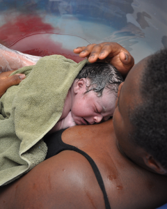 A mother lies back in a birthing pool. She has just given birth and her newborn baby is lying on her chest covered in a green towel