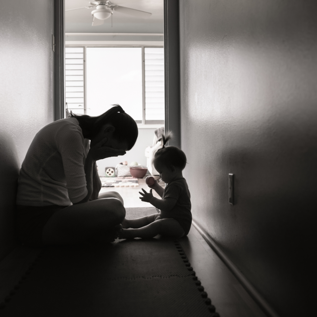 Mother suffering from birth trauma sits in a hallway with her head in her hands whilst her baby plays in front of her